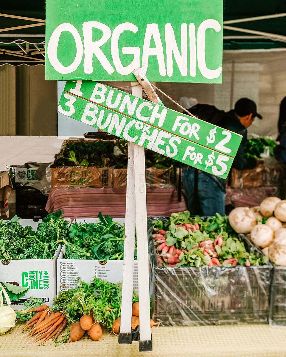 'ORGANIC'이라 쓰인 초록색 팻말 아래 브로콜리, 무, 당근 등 유기농 채소가 상자에 담겨 진열되어 있다.